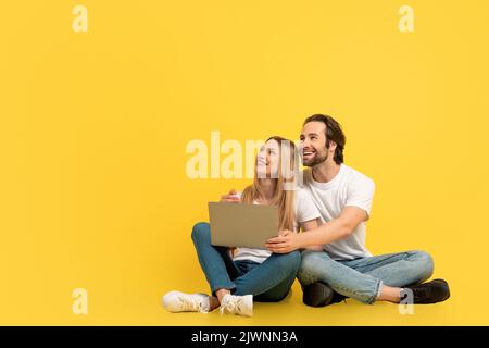 Allegra sorpresa donna e ragazzo caucasico millenario seduto sul pavimento con un notebook e guardando in su lo spazio vuoto Foto Stock