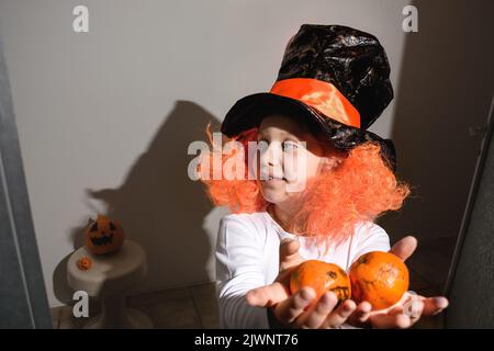 Halloween felice. Una bambina in una parrucca arancione e un cappello nero tiene i tangerini nelle sue mani. Zucca su una sedia. Organizzare una vacanza a casa. Tutte le S Foto Stock