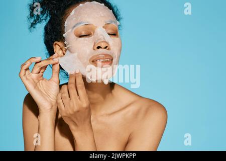 Donna afroamericana applica tessuto cosmetico maschera facciale per idratare, idratare e idratare e brillare la pelle del viso, su sfondo blu. concetto di cura della pelle Foto Stock