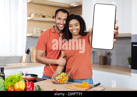 Felice giovane coppia afro-americana nella stessa t-shirt cucinare cibo fatto in casa, mostrare grande smartphone con schermo vuoto Foto Stock