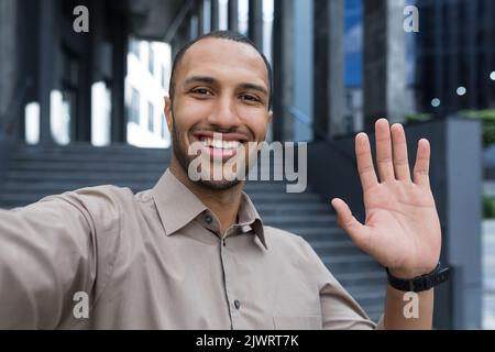 Giovane afroamericano che guarda nella fotocamera dello smartphone, parla in videochiamata usando l'app per telefono, sorridente e felice gesto di saluto, uomo d'affari fuori dal moderno edificio degli uffici. Foto Stock