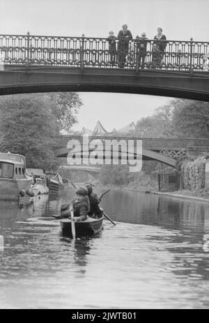 Londra, Inghilterra, circa 1967. Una famiglia che si trova sulla passerella che attraversa il Regent’s Canal al Cumberland Basin guarda i ragazzi del Pirate Club che si addollano una barca sotto di loro. Avanti c'è lo Snowdon Aviary dello Zoo di Londra nel Regent's Park. Il Pirate Club, un club nautico per bambini, è stato fondato nel 1966 presso Gilbey’s Wharf sul Regent’s Canal vicino a Camden, Londra. La loro clubhouse era una vecchia chiatta e un certo numero di piccole barche e canoe erano state donate per l’uso dei bambini. Foto Stock