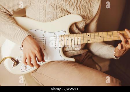Un uomo in un maglione di lana bianca suona abilmente una melodia su una chitarra elettrica bianca. Creazione di musica e creatività. Lezioni di musica. Foto Stock