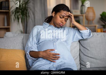 Infelice donna afro-americana incinta sentirsi male seduto a casa Foto Stock