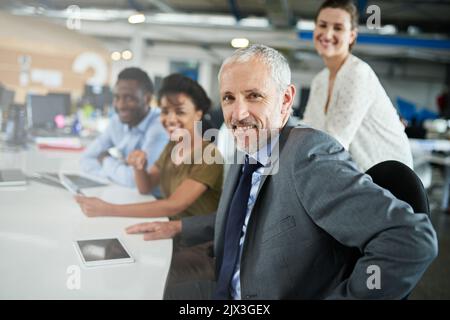 Supportato da una squadra di grandi. Ritratto di un uomo maturo seduto a un tavolo in un ufficio con i colleghi sullo sfondo. Foto Stock