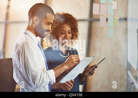 Confronto tra pensieri e positività. Due giovani professionisti che si occupano di brainstorming con note appiccicose su una parete di vetro in un ufficio. Foto Stock