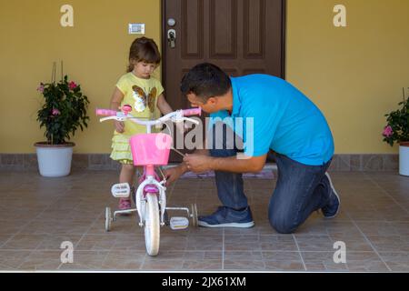 Immagine di un giovane papà con sua figlia nel cortile mentre ripara una piccola bicicletta rosa. Fai da te il lavoro Foto Stock