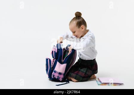 Ragazza carina in uniforme scuola raccoglie uno zaino per le classi isolato su uno sfondo bianco. Foto Stock