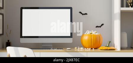 Spazio di lavoro domestico moderno nel tema di Halloween, PC desktop computer bianco schermo mockup, popcorn in zucca secchio su tavolo di legno e bat adesivi neri Foto Stock