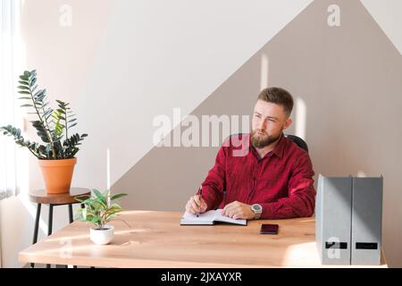 Pianificazione del budget. Uomo pensive barbuto che scrive in un notebook mentre seduto al tavolo in un ufficio moderno in abiti di lavoro, business planning concetto Foto Stock