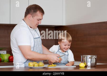 Concetto di cibo e nutrizione. Foto di due allegri papà e figlio che si posano in cucina peeling patate andando a cucinare cibo vegetariano per cena insieme, indossando grembiuli identici Foto Stock