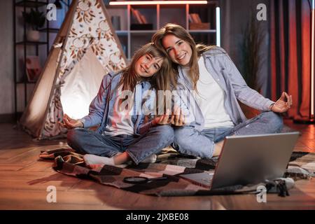 Ragazze caucasiche belle bionde sorelle seduti sul pavimento in posa di lotos fuori wigwam, mentre si utilizza il computer portatile per ascoltare musica rilassante durante il tempo libero sera. Accogliente casa e concetto di famiglia. Foto Stock