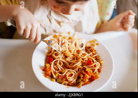 Bambino che mangia spaghetti con verdure. Bambino che mangia divertente. Ragazzo dai capelli castani con il viso coperto di salsa. Fine settimana, ambiente caldo e accogliente in cucina Foto Stock