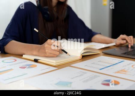 Una giovane donna asiatica lavora da casa in cucina prendendo appunti mentre si utilizza un computer portatile per il remoto. Foto Stock