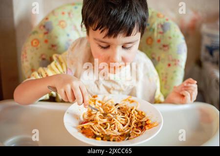 Bambino che mangia spaghetti con verdure. Bambino che mangia divertente. Ragazzo dai capelli castani con il viso coperto di salsa. Fine settimana, ambiente caldo e accogliente in cucina Foto Stock