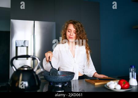 Giovane donna caucasica sorridente cuoce la colazione su una padella. Giovane donna adulta che fa omelette su una stufa a gas. Ragazza che prepara il suo pasto su un Foto Stock