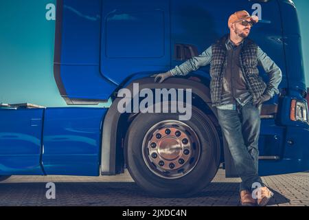 Trucker di mezza età caucasico orgogliosamente in piedi davanti al suo camion semi-rimorchio blu grande che guarda alla sinistra in attesa del carico. Tran. Per impieghi pesanti Foto Stock