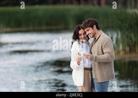 Coppia allegra in cardigan lavorato a maglia che brinda vino vicino fiume sfocato, immagine stock Foto Stock