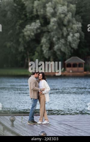 Donna allegra abbracciando ragazzo in cardigan e tenendo vino vicino al lago, immagine stock Foto Stock