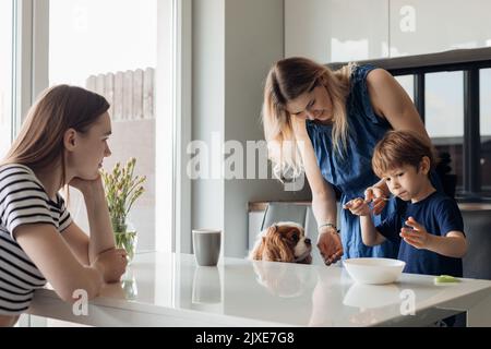 Due donne calme e deluse e un ragazzo poco triste, rifiutando il cibo siedono con il cane in cucina. Mangiatore picky, abitudini differenti Foto Stock