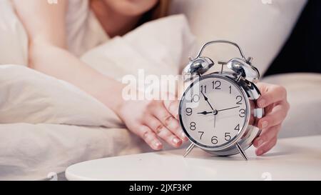 Vista ritagliata di una donna sfocata che tocca la sveglia in camera da letto al mattino, immagine stock Foto Stock