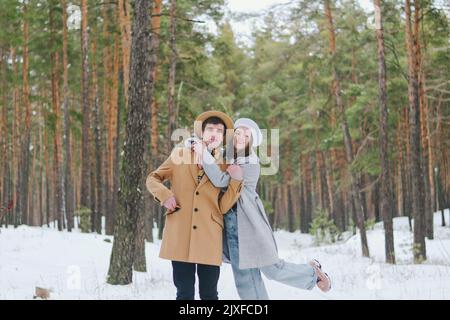 divertente famiglia, coppia in parco nevoso, foresta. Giovane donna, ragazza e uomo in cappello camminando vicino a pini innevati. Bere caffè. Divertendoti. Famiglia Foto Stock