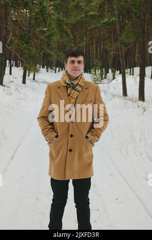 Ritratto di giovane divertente in jeans, cappello e giacca in parco nevoso, foresta. Camminare vicino a pini innevati. Divertendoti. Vacanze invernali in famiglia. Foto Stock