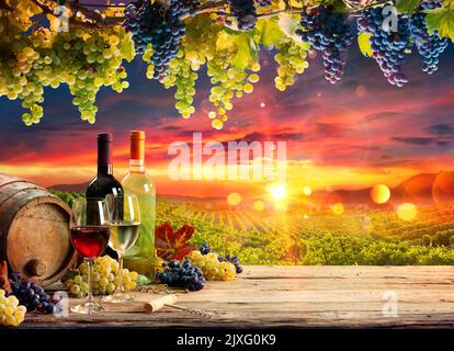 Bicchieri da vino e bottiglia con uva e barile in Vigneto al tramonto Foto Stock