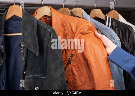 Uomo controlla giacca in pelle arancione in un negozio di seconda mano. Varie giacche d'epoca appendono su una rastrelliera di abbigliamento. Thrifting e sostenibilità in abbigliamento conce Foto Stock