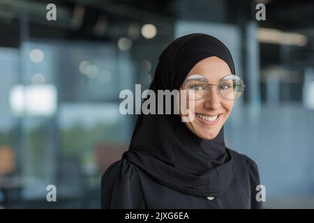 Primo piano ritratto di una donna d'affari musulmana in hijab sorridendo e guardando la macchina fotografica, lavoratrice in occhiali che lavorano all'interno di un moderno edificio di uffici. Foto Stock