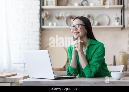 Blog. Freelance. Una giovane donna in occhiali seduti a casa in cucina con un computer portatile, scrivendo un post, lavorando sui social network, chattando. Ha piegato le mani sotto il mento, pensando, sorridendo. Foto Stock