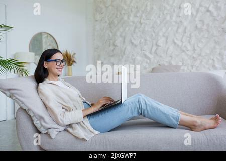 Lavoro online. Giovane donna d'affari in occhiali, freelance che lavora a casa con un notebook. Stenditi comodamente sul divano in jeans e una camicia, digitando Foto Stock