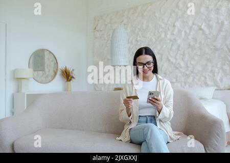 Shopping online. Soddisfatto giovane bella donna in occhiali seduti sul divano a casa. Tiene una carta di credito e telefono, paga il prodotto, consegna ordini. Foto Stock