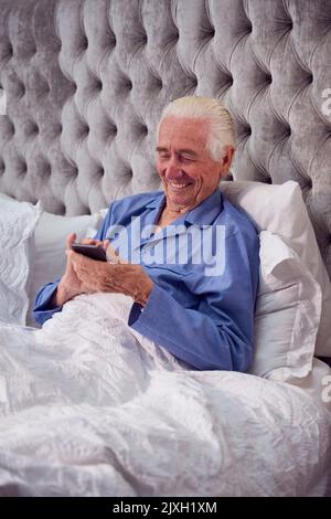Sorridente uomo anziano a casa con pigiama a letto utilizzando il telefono cellulare Foto Stock