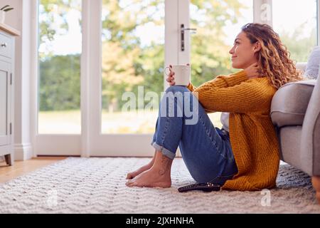 Giovane donna in intimo caldo Jumper seduto sul pavimento a casa guardando fuori dalla finestra con bevanda calda Foto Stock