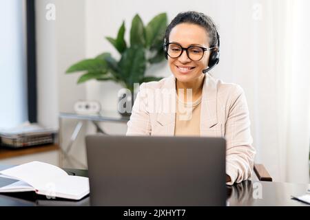 Sorridente dipendente di un call center che utilizza un auricolare e un computer portatile per comunicare online con i clienti, donna che parla nel microfono e digita sulla tastiera, assegni e segni Foto Stock