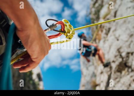 Primo piano di un dispositivo di assicurazione con un ragazzo sulla parete di arrampicata scogliera. Appeso su una corda in un'imbracatura da arrampicata e il suo compagno lo cinge sul gallo Foto Stock