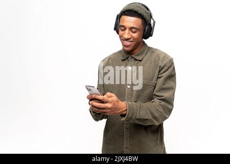 Divertente uomo multirazziale che indossa le cuffie con uno smartphone, trascorre il tempo libero online, guarda video, ascolta musica e sorrisi, felice uomo con auricolari tiene il telefono, isolato su bianco Foto Stock
