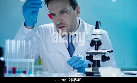 un attento scienziato che guarda la provetta con campione rosso vicino al microscopio in laboratorio, immagine stock Foto Stock