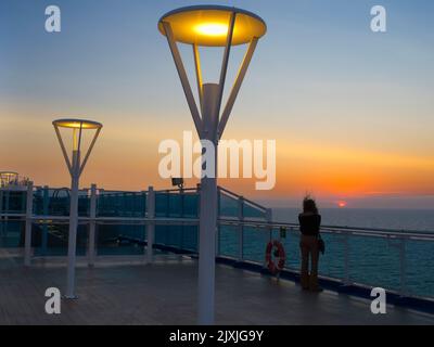 Scena tranquilla; una donna con i capelli che volano al vento guarda un tramonto drammatico sul Mare del Nord. Siamo a bordo di una nave da crociera diretta verso il gr Foto Stock