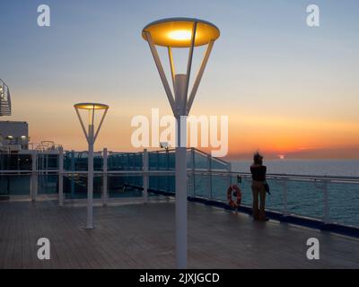 Scena tranquilla; una donna con i capelli che volano al vento guarda un tramonto drammatico sul Mare del Nord. Siamo a bordo di una nave da crociera diretta verso il gr Foto Stock