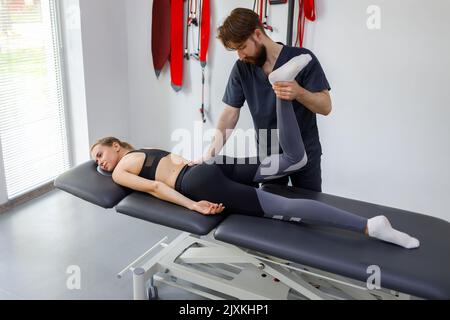 Fisioterapista maschile che fa il trattamento di gambe e ginocchia su una giovane paziente in posizione supina. Donna atletica in visita la sua riabilitazione specialis Foto Stock