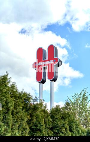 RIGA, LETTONIA - 13 MAGGIO 2022: Logo del ristorante Hesburger rosso contro il cielo nuvoloso blu durante la giornata di sole. Verde alberi di conifere sotto. Foto Stock