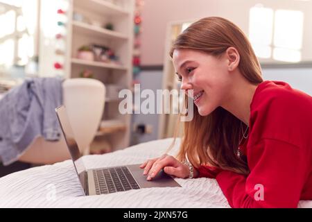 Ragazza adolescente che indossa pigiama utilizzando notebook sdraiato sul letto in camera da letto Foto Stock