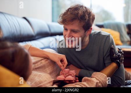 Uomo mettere le scarpe di bambino sullo stomaco di partner incinta con braccio protesico a casa Foto Stock