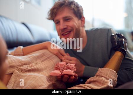 Uomo mettere le scarpe di bambino sullo stomaco di partner incinta con braccio protesico a casa Foto Stock