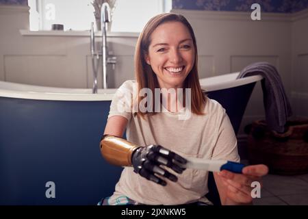 Ritratto di donna eccitata con braccio protesico seduto sul pavimento del bagno con test di gravidanza positivo Foto Stock