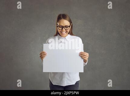 Giovane bella donna felice si alza su uno sfondo grigio e tiene un bianco foglio nelle sue mani. Foto Stock