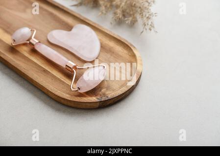 Rullo massaggiante viso in pietra di quarzo rosa e massaggiatore gua sha in pietra naturale su fondo di cemento chiaro, vista dall'alto Foto Stock