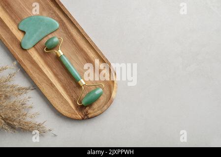 Rulli per massaggio viso in giada e massaggiatore gua sha in pietra naturale in giada su fondo in cemento chiaro, vista dall'alto Foto Stock